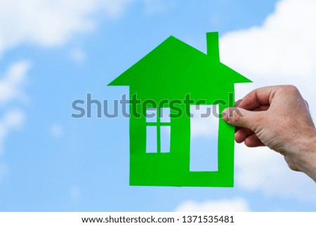 Green paper house in hand on the background of blue sky.  Hand holding house paper with blue sky background. Concept of home, real estate, insurance.