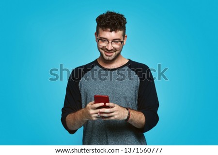 Cheerful young hipster boy with eyeglasses holding smartphone and and writing message on cell phone over blue background.