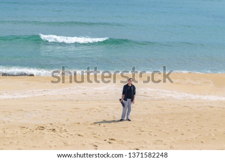 Young man photographer with tripod in hand standing in the middle of sandy beach with sea and waves on background. Travel photographer concept. Copy space, horizontal image.