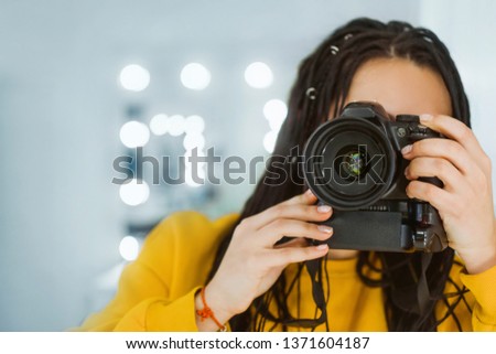 a young girl photographer, holding a camera in her hands and taking pictures against the background of lights, she is wearing a yellow jacket and with dreadlocks, focus on the camera.
