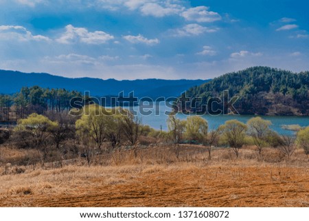 Landscape of field in countryside with lake and mountains in background under blue partly cloudy sky. 
