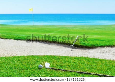 Golf stick and ball on grass against the sea. Rake near the sand.