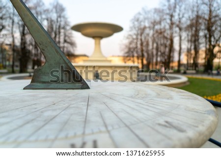 Sundial with a blurred fountain, trees and a garden in the background. During sunset