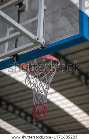 An interior space of a basket ball court showing a scaffolding metal roof sheet with a framework of metal truss with sunlight goes through and a basketball ring as a focal point.