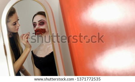 Halloween. Make-up in horror style. The view in the mirror of a professional makeup artist makes the model's bloodied mouth for a photo shoot. Red background. Light spots.