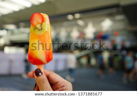 Melting ice cream in a hot summer day, kiwi fruit strawberry and tangerine sorbet stick in hand of woman with dark brown painted fingernails on blurred background of street market. (selective focus)