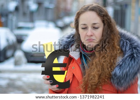 Exaggeratedly sad red-haired girl in warm clothes in winter with a euro sign and an arrow down. Concept - the euro falls