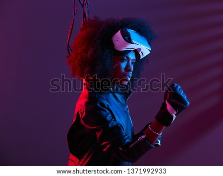 Stylish curly dark haired girl dressed in black leather jacket and gloves is wearing the virtual reality glasses on her head in the dark studio with neon light