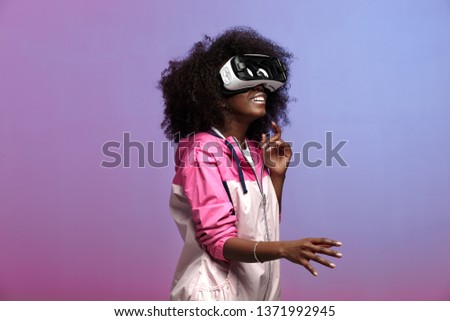 Mod young brown-haired curly girl dressed in the pink sports jacket uses the virtual reality glasses in the studio on neon background