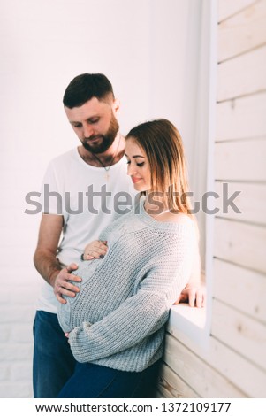 Young pregnant couple near the window of the house, on the background of a brick white wall and a wooden window sill