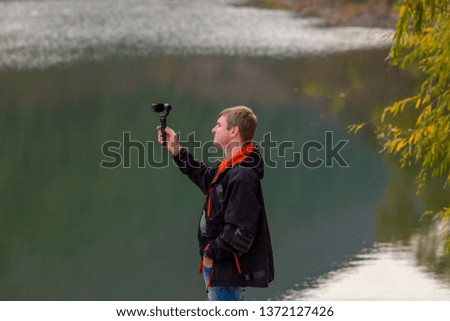 A man takes an action camera. A blond man in a black jacket and jeans holds a video camera and takes pictures. The guy stands at the lake , in which the branches of the willow bend