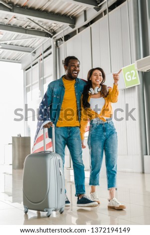 smiling african american couple with american flag and suitcase walking  in departure lounge in airport 