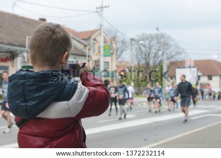 Group of runners dressed in black are passing by a kid photographer during a Belgrade marathon race. Boy is trying to capture a perfect moment with his digital camera.