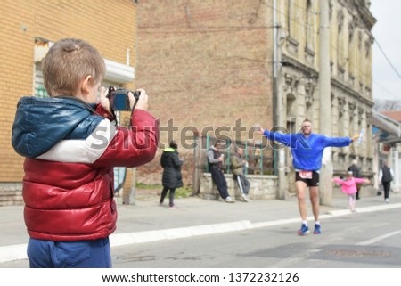 A small boy photographer is photographing a marathon race. He holds his digital camera while runners are passing by. One runner is posing for him.