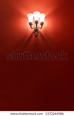 Lantern with three lamps on the red wall. From the lamp down the shadows fall on the wall. The wall is dark red. At the bottom of the photo there is a large space for text