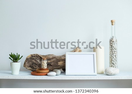 Modern room decoration with picture frame mockup. Shelf against white wall with decorative candle, glass, wood and rocks. Home plant in pot.