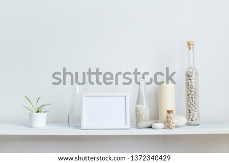 Modern room decoration with picture frame mockup. Shelf against white wall with decorative candle, glass and rocks. Potted spider plant.