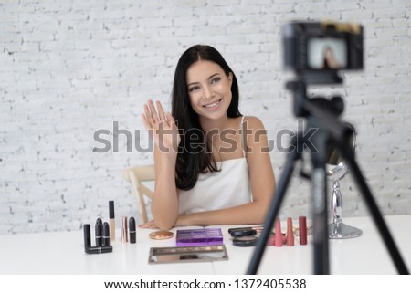 Portrait of beautiful young woman professional beauty vlogger or blogger saying hi and waving with recording video sitting on white brick wall background.Blogger beauty business concept.Making Vlog.