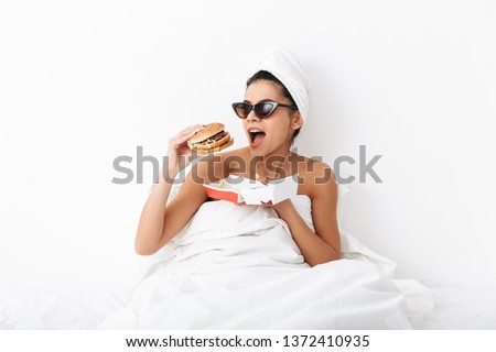 Image of a beautiful emotional woman with towel on head lies in bed under blanket isolated over white wall background wearing sunglasses eat burger.