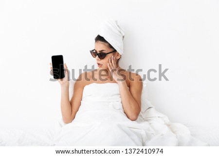 Image of a beautiful surprised emotional woman with towel on head lies in bed under blanket isolated over white wall background wearing sunglasses showing display of phone.