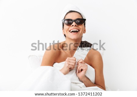 Image of a beautiful woman with towel on head lies in bed under blanket isolated over white wall background wearing sunglasses eat chocolate.