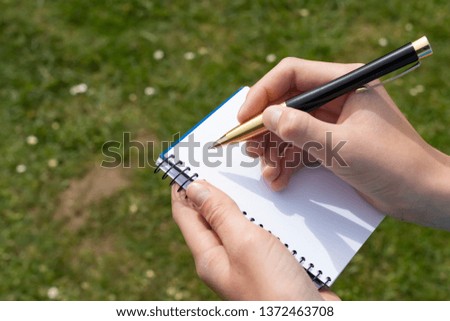 Boy's hand with a yellow-black pencil over an open notepad on a background of green grass and white flowers in the park. Clean white sheet. Going to write or draw a picture.