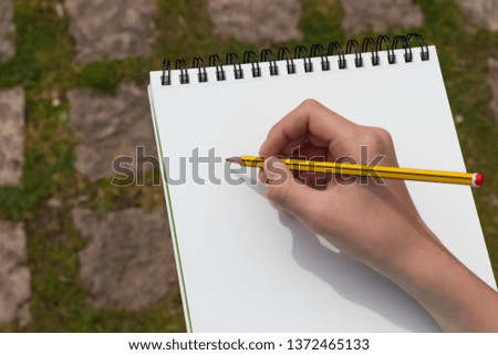 The boy's hand with a yellow-black pencil over an open notepad on the background of paving stones in the park. Clean white sheet. Going to write or draw a picture. The concept of life planning.