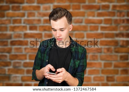 White young guy writes a message on mobile phone and smiles. Man on brick wall background