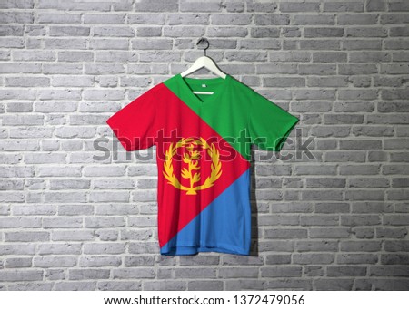 Eritrea flag on shirt and hanging on the wall with brick pattern wallpaper. Red triangle on blue and green triangle with olive branch on gold color. 