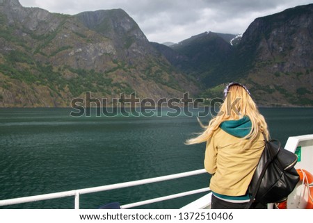 Young woman with a backpack on the deck of the ferry looks at the Norwegian fjords. Woman blonde tourist on a ship admires the nature of Norway. A young woman travels alone. Tourist concept.