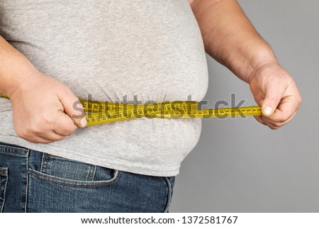 A man measures his fat belly with a measuring tape. on a gray background Royalty-Free Stock Photo #1372581767