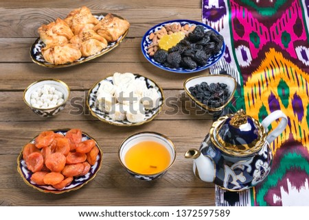Traditional Uzbek sweets - dried apricot, rohat Turkish delight, raisin, samsa, almond, teapot with tea and bowl with national ornament on wooden table. Flat lay.