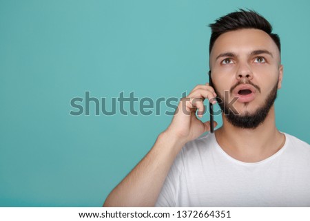 Shocked young bearded hipster in white t-shirt using phone and talking with bright smile. Copy space.