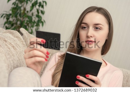 Attractive woman uses tablet, holding a plastic card, sitting at home on the couch, toned