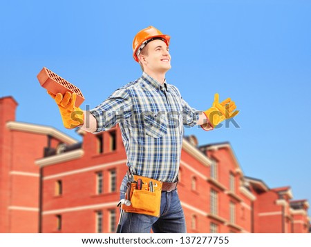 Happy manual worker holding a brick and spreading arms with a building in a background