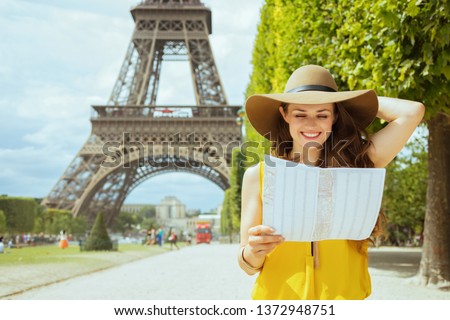 happy young solo tourist woman in yellow blouse and hat at Champ de Mars overlooking Eiffel tower in Paris, France looking at the map.