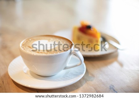 White cappuccino cup with with latte art heart and cake on light brown wood background lit by bright morning sunlight. Slow living concept