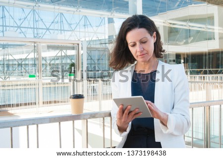 Medium shot of serious middle-aged Caucasian woman in white jacket standing in office building, holding tablet in hands, typing. Work, communication concept