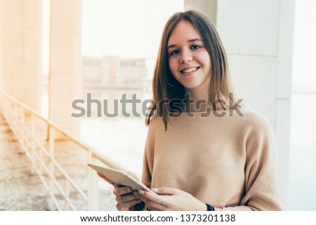 Smiling female teenager browsing on tablet computer outdoors. Pretty young woman wearing casual clothes and standing with railing and lake in background. Education and technology concept. Front view.