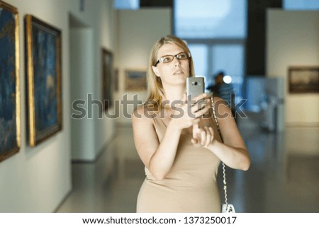 Female visitor wearing glasses looking at exhibits and taking picture on smartphone in art museum
