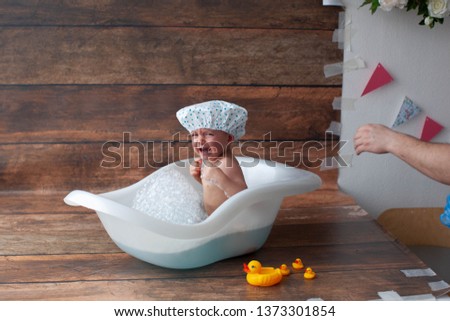 Baby boy takes bath with yellow ducks and watering can.  Air bubbles and bath foam. One year old boy in swimming cap. Little boy crying. He does not like to swim. Bathroom.