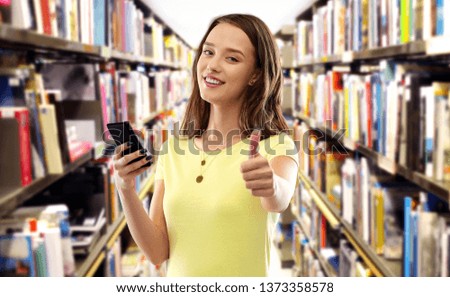 technology, education and people concept - smiling young woman or teenage girl in blank yellow t-shirt with smartphone showing thumbs up over book shelves in library background