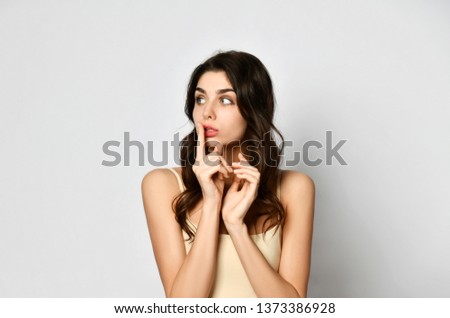 A bit scared beautiful woman holds her finger at her mouth and make shhh like asks to speak softly, keep silence or a secret on gray background