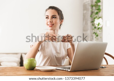Image of a beautiful young student girl with apple sitting indoors using laptop computer drinking coffee.