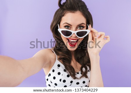 Portrait of stylish pin-up woman 20s in retro sunglasses smiling while taking selfie photo isolated over violet background