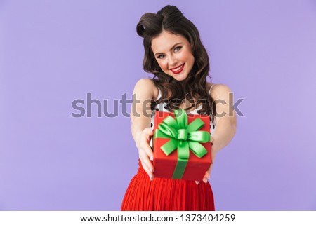 Portrait of brunette pin-up woman 20s in retro polka dot dress rejoicing while holding birthday present box isolated over violet background