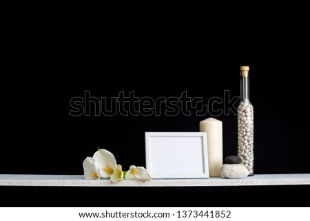 Modern room decoration with picture frame mockup. Shelf against black wall with decorative candle, glass, wood and rocks. Home plant calla.