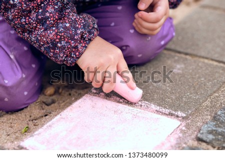 A young girl of about three years painting on the concrete floor of a playground with some children's street chalk. Seen in Nuremberg, Germany, in March 2019