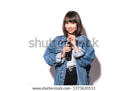 Picture of young woman in jeans jacket drinking soda standing isolated over grey wall background.