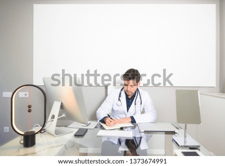 Doctor sitting at the desk of his clinic office writing on his paper agenda. White blank poster with copy space in the background.